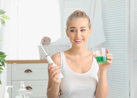 Woman holding glass of green mouthwash in one hand and electric toothbrush in the other