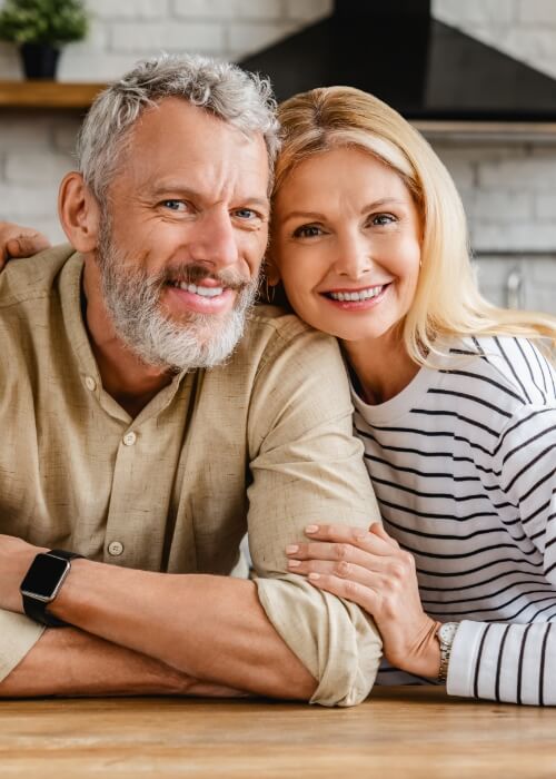 Older man and woman smiling in their kitchen after receiving dental services in Cumming